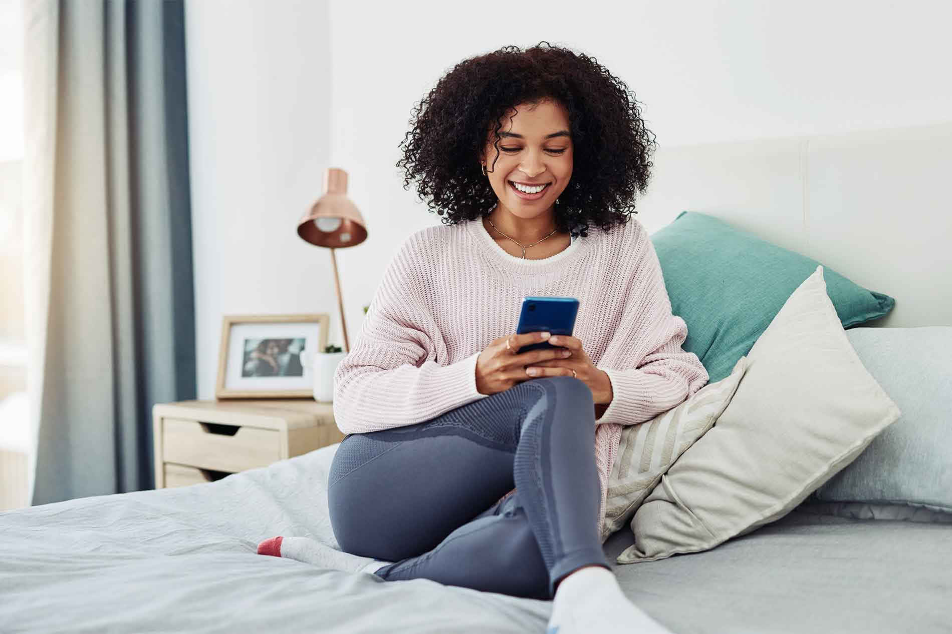 Happy young woman looking at phone on bed