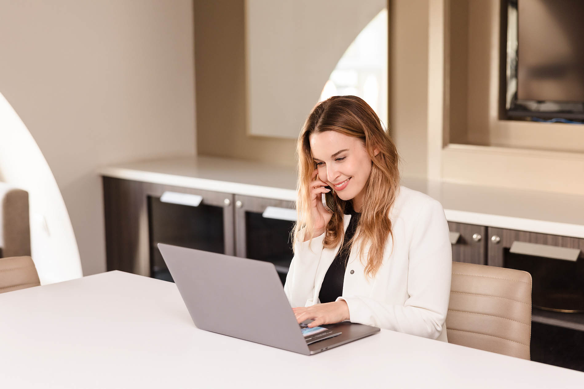 A woman sits at a table in an apartment community, using a laptop and talking on the phone